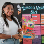 Nillyreth sostiene productos de su negocio comunitario frente a un cartel de emprendimiento.